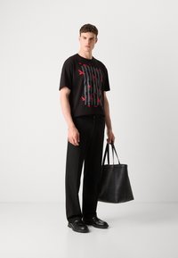 Schwarzes T-Shirt mit rotem Vogelmotiv, schwarze Hose und schwarze Ledertasche, stehend vor einem hellgrauen Hintergrund.