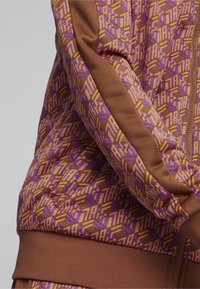 Veste de sport marron avec un motif géométrique violet et jaune, dotée d'un poignet côtelé et d'une bande latérale, fabriquée en tissu lisse et extensible.