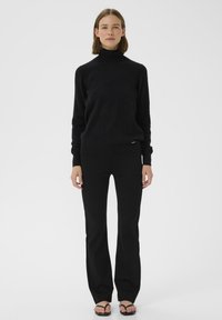 Maglione a collo alto nero con una texture morbida, abbinato a pantaloni neri a campana. Entrambi gli articoli presentano un design minimalista, creando un aspetto coerente.