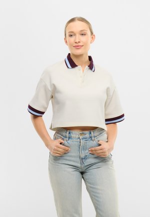 Joven con un polo corto color crema con franjas moradas y azul claro en el cuello y las mangas, combinado con unos jeans de tiro alto en azul claro.