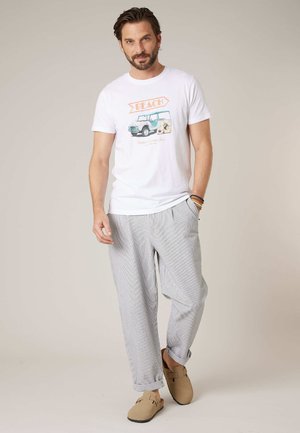 Homme debout portant un t-shirt blanc avec un motif de plage, un pantalon gris clair, des chaussures beige à enfiler et des bracelets au poignet droit, sur un fond neutre.