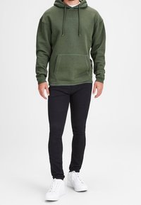 Olivgrüner Kapuzenpullover mit Kängurutasche, Zugbändern und dezentem Logo. Kombiniert mit eng anliegenden schwarzen Jeans und weißen Turnschuhen.