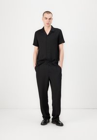 Chemise noire à manches courtes avec un devant à boutons et une texture subtile, assortie à un pantalon noir à coupe décontractée et avec des plis.
