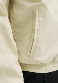 Veste beige clair en tissu avec une finition texturée. Équipée d'une poche latérale avec un détail de couture subtil et d'une taille élastique.
