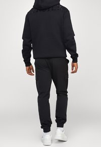 Hoodie noir avec un design texturé au dos, associé à un pantalon cargo noir. La tenue comprend des poignets élastiques et des baskets blanches.