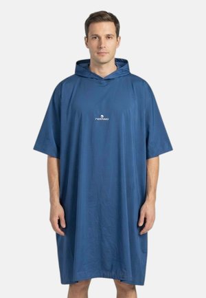 Uomo che indossa un poncho blu largo con cappuccio e maniche corte, con un piccolo logo bianco sul petto, in piedi davanti a uno sfondo semplice.