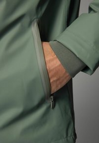 Jacheta verde impermeabilă, cu o textură netedă, având un manșon ajustat și un buzunar lateral, evidențiind un design elegant și modern.