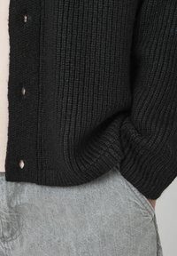 Cardigan en tricot noir avec une texture côtelée et un design à boutons, porté sur une chemise claire, associé à un pantalon en tissu gris.
