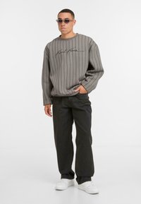 Uomo che indossa un felpa grigia a righe con scritta, jeans neri, sneakers bianche e occhiali da sole, in piedi con una mano in tasca.