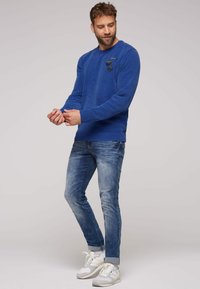 Maglione blu con texture, con polsini e orlo a coste, caratterizzato da tre toppe decorative sul petto, abbinato a jeans blu sbiaditi e scarpe da ginnastica bianche.