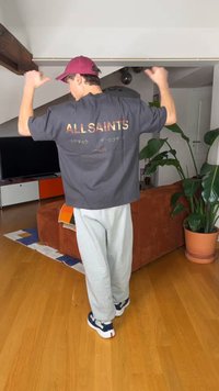 Mees, kes kannab burgundia värvi mütsi ja halli AllSaints T-särki, paindes oma käsi elutoas, kus on taimi ja puidust põrand.