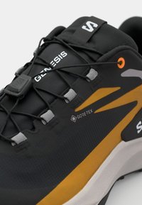 Chaussure de sport noire et grise avec une tige texturée, des accents orange et jaune, et une étiquette GORE-TEX. Lacets élastiques avec un mécanisme de verrouillage.