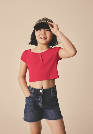 Ung pige smilende, iført en rød cropped skjorte, denimshorts og leopardmønstret kasket, stående mod en beige baggrund.