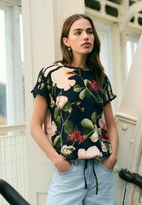 Květinová blůza s námořnickým pozadím, krátkými rukávy se zoubkovanými okraji a velkými růžovými a zelenými květinovými vzory. Kombinováno se světle modrými džíny.