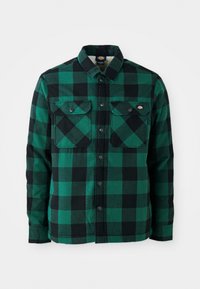 Chemise en flanelle à carreaux verts et noirs avec un col à bouton, deux poches poitrine et des fermetures à pression en métal noir. Texture douce.