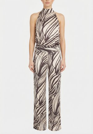 Naine kannab varrukateta kõrge kaelusega kombinesooni mustade ja beežide abstraktsete sebratriipudega, seisab ühevärvilise valge tausta ees.