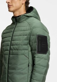 Uomo che indossa una giacca a vento verde trapuntata con cappuccio e una tasca con zip nera cucita sulla manica sinistra superiore.