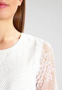 Bílá blůza s texturovanými krajkovými rukávy. Hlavní část má hladký povrch, zatímco rukávy ukazují květinové a tečkované vzory.