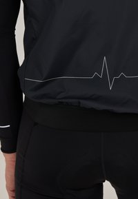 Schwarze Fahrradjacke mit einem Herzschlag-Linien-Grafik, kombiniert mit schwarzen, enganliegenden Shorts mit strukturiertem Bund und gepolstertem Sitz.