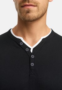 Sort henley-t-shirt med korte ærmer og hvid krave. Har fem mørke knapper og en blød, glat tekstur. Nærbillede af halsudskæringen.