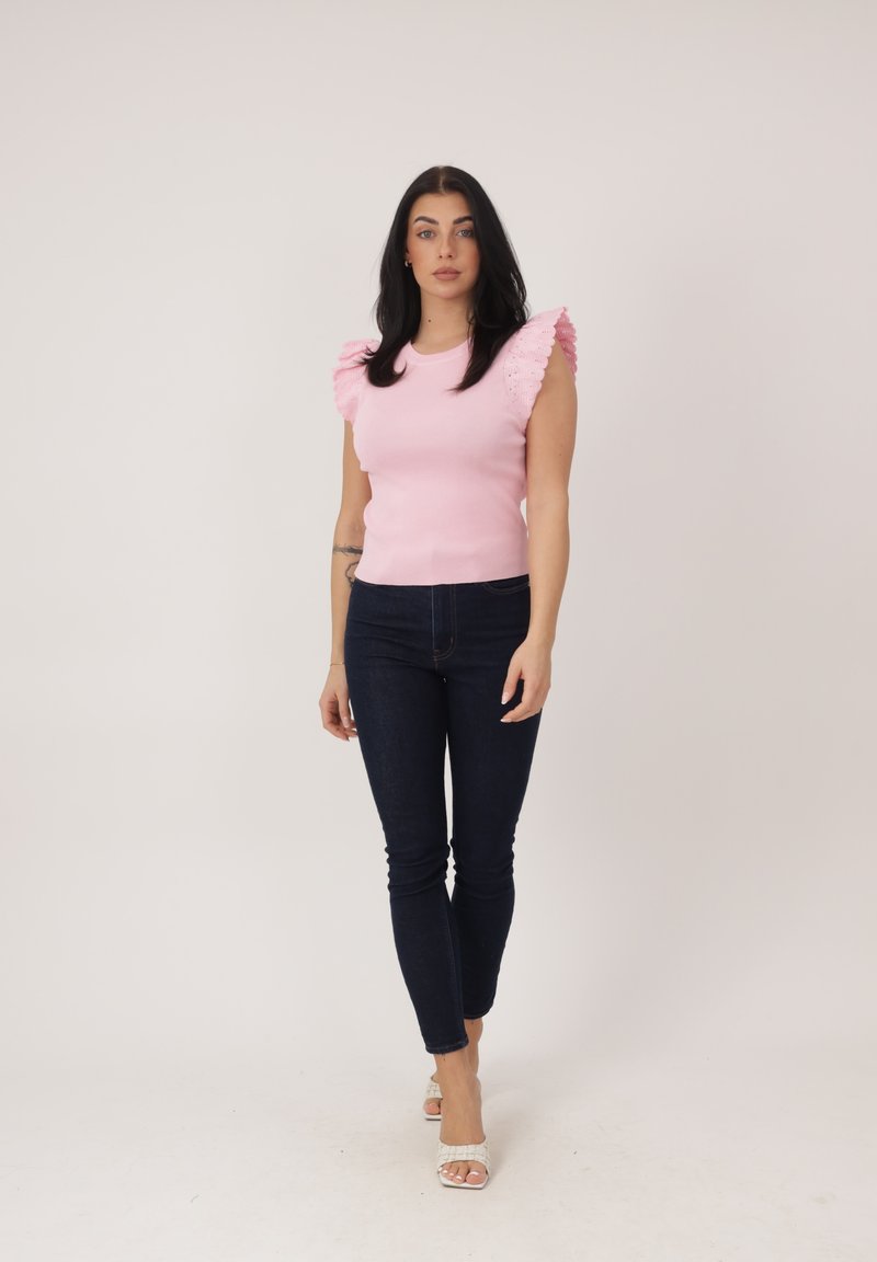 Frau mit schwarzen Haaren, die ein pinkfarbenes ärmelloses Oberteil mit Rüschen an den Schultern, dunkle Skinny-Jeans und weiße Sandalen mit Absatz trägt und vor einem einfarbigen Hintergrund steht.