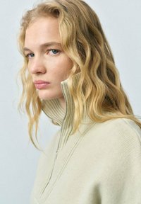 Light beige sweater with a high collar and zip closure. The fabric appears soft and ribbed, with textured accents along the shoulders.