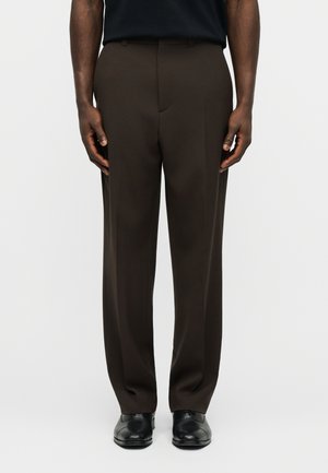 Άνδρας που φορεί καφέ σκουρό χρώμα παντελόνι με ίσια γραμμή, μαύρα επίσημα παπούτσια και μαύρο πουκάμισο, στέκεται μπροστά σε απλό φωτεινό φόντο.