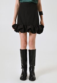 Gonna nera con orlo a volant, realizzata in tessuto morbido. Abbinata a stivali neri alti fino al ginocchio con texture lucida e un piccolo dettaglio dorato.