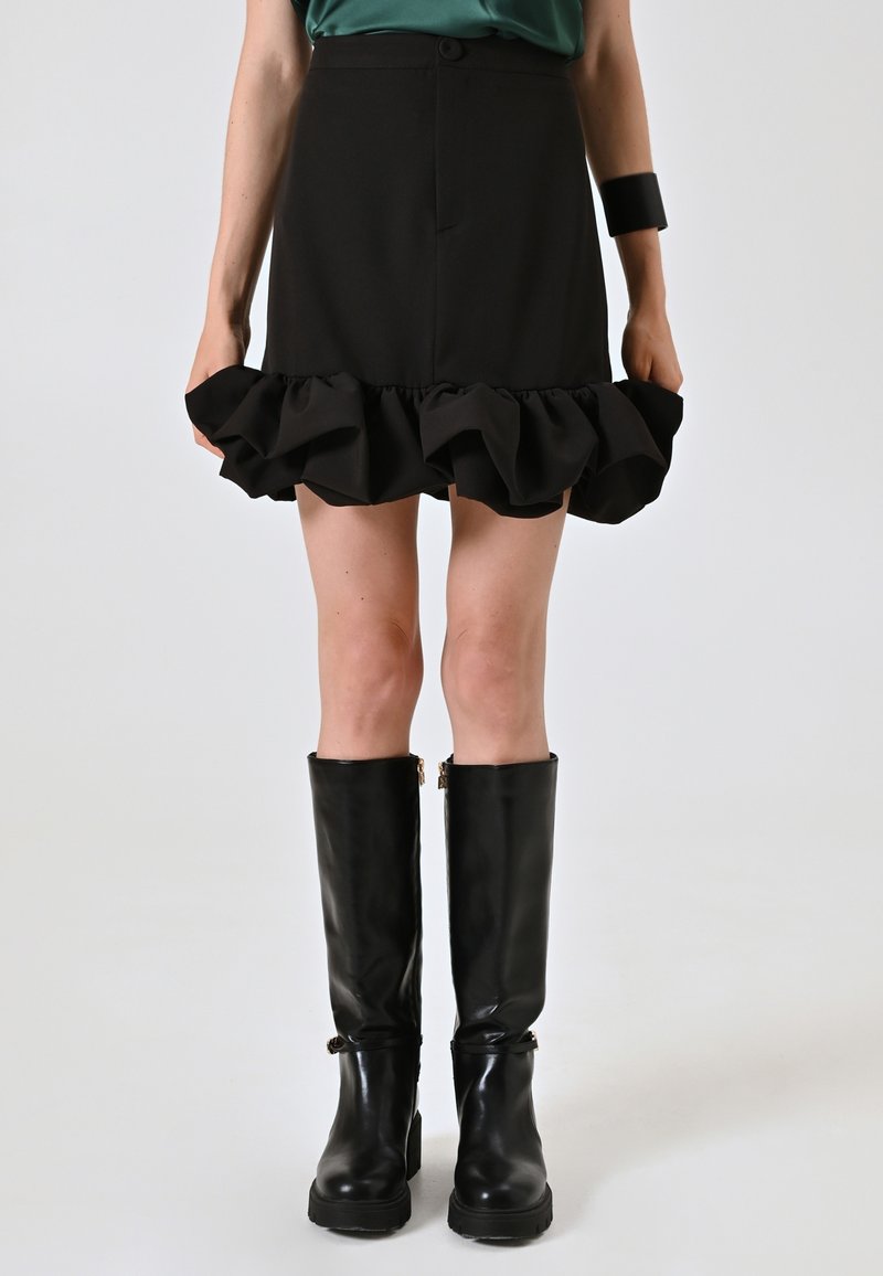 Gonna nera con orlo a volant, realizzata in tessuto morbido. Abbinata a stivali neri alti fino al ginocchio con texture lucida e un piccolo dettaglio dorato.
