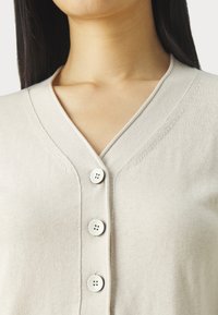Beżowy kardigan w serek z czterema okrągłymi guzikami, o miękkiej teksturze i delikatnym wzorze dzianiny. Widoczne detale na kołnierzu i listwie.
