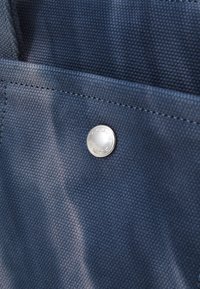 Marineblauwe stoffen tas met een gestructureerd oppervlak, voorzien van een zilveren knoopsluiting met het merkembleem, en versterkte stiksels langs de randen.