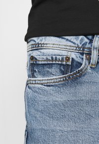 Blå denimjeans med femficksdesign, kontrasterande gula sömmar, en sliten textur och detaljer av kopparnitar.