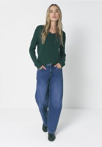 Tmavě zelený svetr s dlouhým rukávem a výstřihem do V, doplněný širokými modrými džíny. Modelka stojí s rukama v kapsách a má na sobě černé boty.