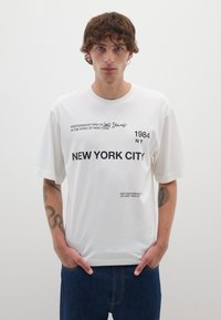 Camiseta de algodón blanca con mangas cortas. Presenta texto negro: "NUEVA YORK" y varios lugares y fechas. Corte casual, cuello redondo.