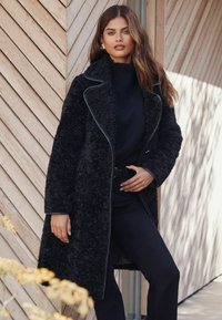 Palton lung negru texturat cu guler tip rever și bordură contrastantă, purtat peste un pulover negru tricotat și pantaloni mulați, pe fundal de lemn.