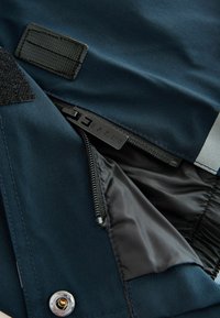 Marineblaue Jacke mit halb offenem Reißverschluss, der das schwarze Innenfutter zeigt, mit "BAKER" eingraviert am Reißverschlussanhänger und einem Metall-Druckknopf darunter.
