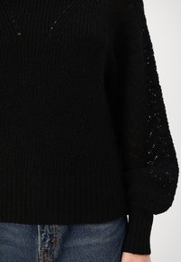 Maglione nero lavorato a maglia con orlo a costine, motivo testurizzato e dettagli in pizzo sulle maniche. Abbinato a jeans di denim.