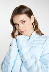 Giacca a vento trapuntata azzurro chiaro con chiusura a zip e maniche ampie. Tessuto liscio con motivi di cuciture diagonali e design minimalista.