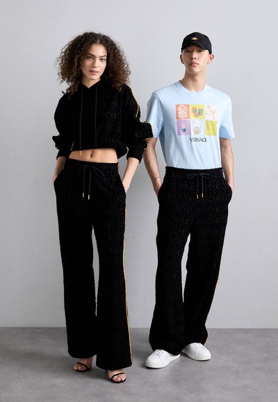Hoodie court en velours noir avec des accents jaunes et un pantalon large assorti ; le modèle masculin porte un t-shirt graphique bleu clair et des baskets blanches.