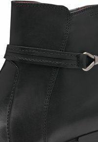 Bottine en cuir noir avec une texture lisse, des coutures décoratives et une sangle argentée autour de la cheville.