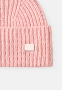 Rózsaszín bordás kötött sapka, puha gyapjúból készült, elöl egy téglalap alakú fehér címkével, amelyen a "TOMMY JEANS" felirat látható.
