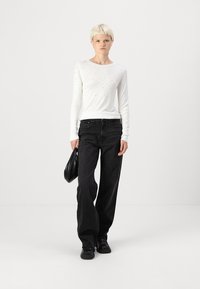 Top alb cu maneci lungi si mici accesorii argintii, asortat cu blugi negri cu talie inalta si croi larg, incaltat cu pantofi negri si tinand o geanta neagra.