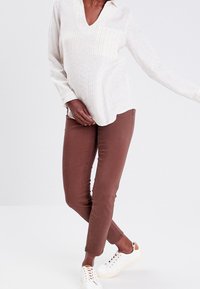 Blouse à manches longues, crème, avec des rayures verticales et une poche, associée à un jean skinny marron et des baskets blanches avec des accents beiges.