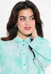 Camicia button-up turchese chiaro con colletto, maniche lunghe a sbuffo e bottoni turchesi lucidi; il tessuto sembra morbido e leggermente strutturato.