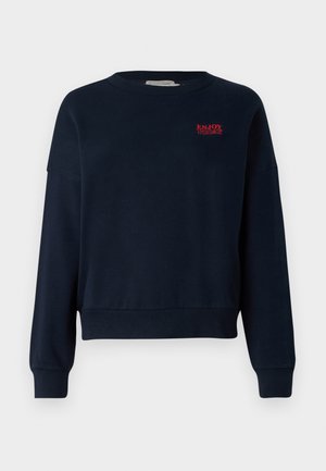 Navy zila sporta krekls ar brīvu piegriezumu, apaļu kakla izgriezumu un ribota piedurknes. Krūtīs ir sarkanas izšūtas uzraksts "IZBAUDI ŠODIEN".