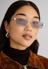 Ochelari de soare rectangulari cu lentile colorate, rame subțiri din argint și un design minimalist. Modelul poartă un pulover negru cu guler înalt și o jachetă din nubuc maro.