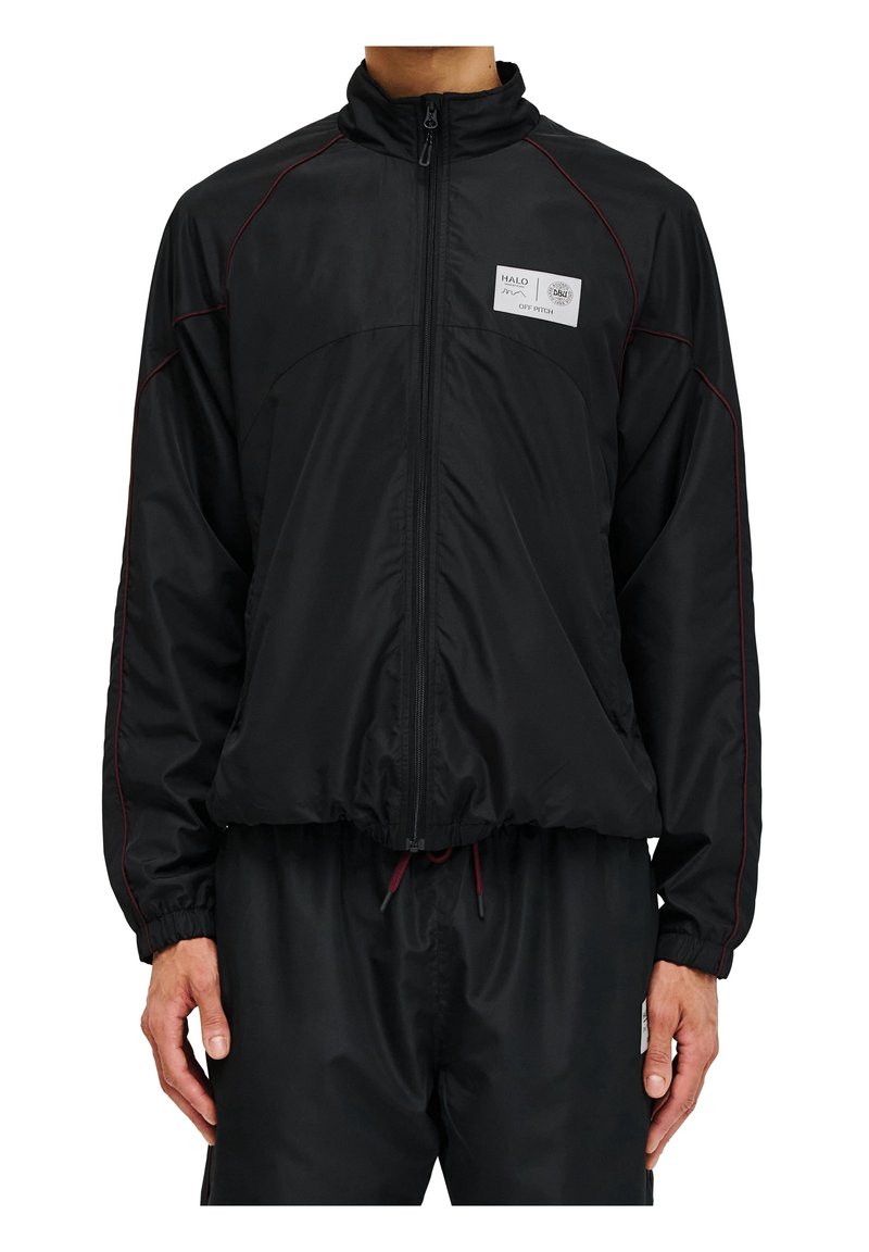 Homme portant un coupe-vent noir avec fermeture éclair et un pantalon assorti, la veste comporte un petit patch blanc rectangulaire sur la poitrine gauche avec du texte.