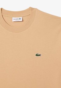 T-shirt in cotone beige con scollo rotondo, caratterizzato da un piccolo logo di coccodrillo verde sul lato sinistro del petto. Tessuto morbido, design dalla vestibilità classica.