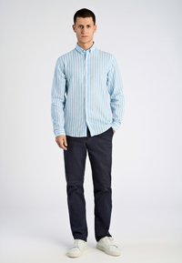 Camicia a righe blu chiaro e crema con maniche lunghe, abbinata a pantaloni scuri e sneaker bianche, in piedi davanti a uno sfondo neutro.