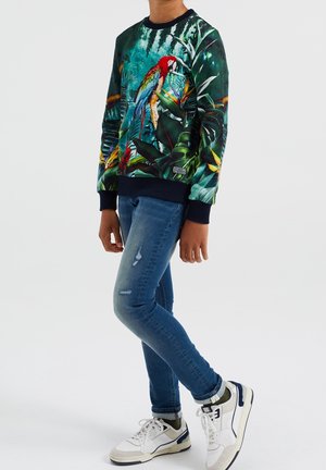 Sweatshirt avec un imprimé de perroquet sur un fond vert foncé, avec des poignets et un ourlet en côtes. Porté avec un jean bleu déchiré et des baskets blanches.